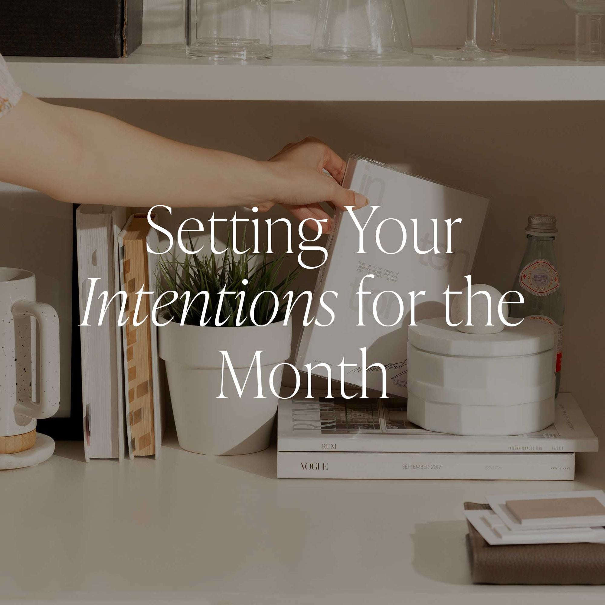An image of a hand reaching for a notebook on a white shelf filled with books, indoor cactus, and a white decorative ceramic jar. It has a text overlay that reads: "Setting Your Intentions for the Month."