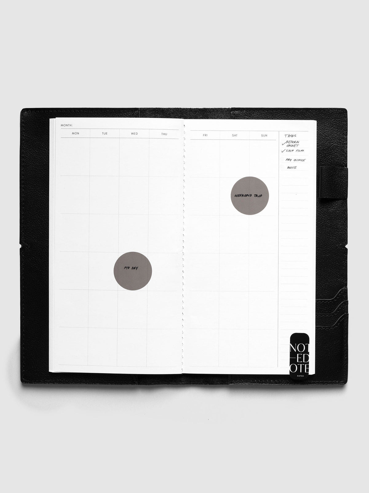 Monthly Overview A5 Slim calendar spread inside a black travel notebook cover, with PTO Day and Weekend Trip circle stickers, a task list and a Noted sticker
