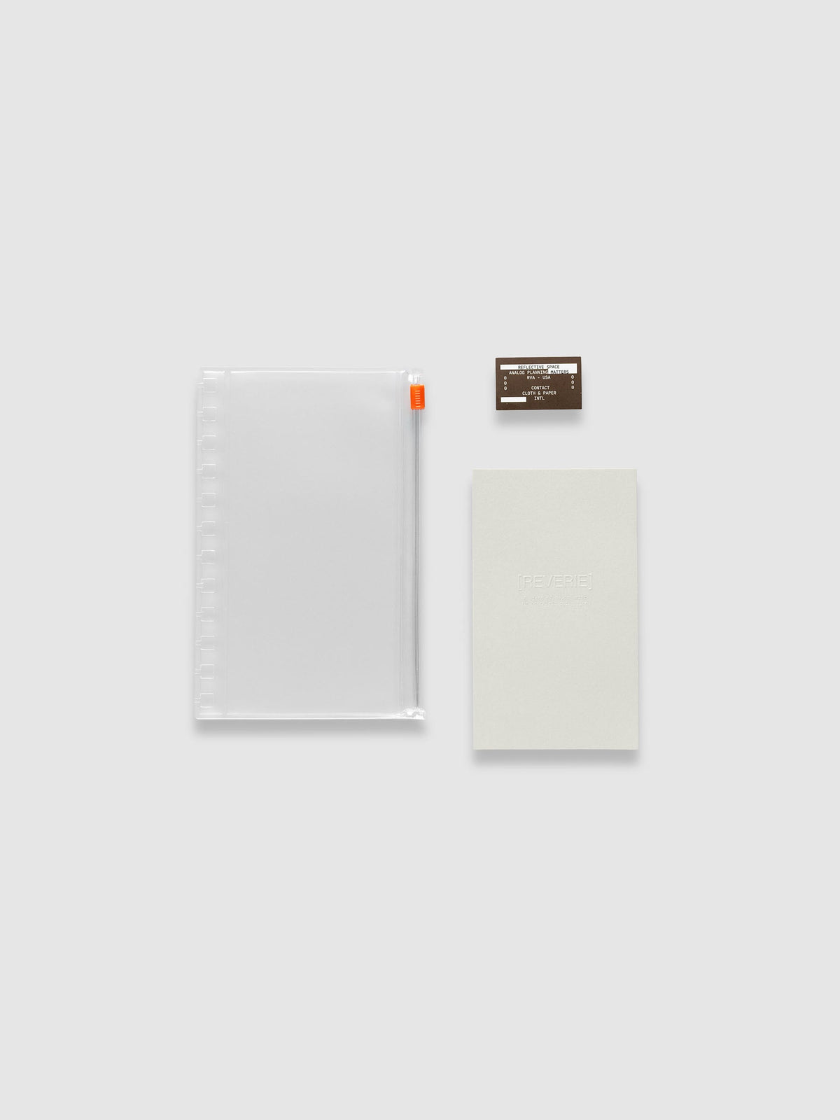 One clear zippered pouch with an orange zipper pull, one light grey embossed paper dashboard and one brown sticker laid side by side. Shown in size CP Petite Spiral.