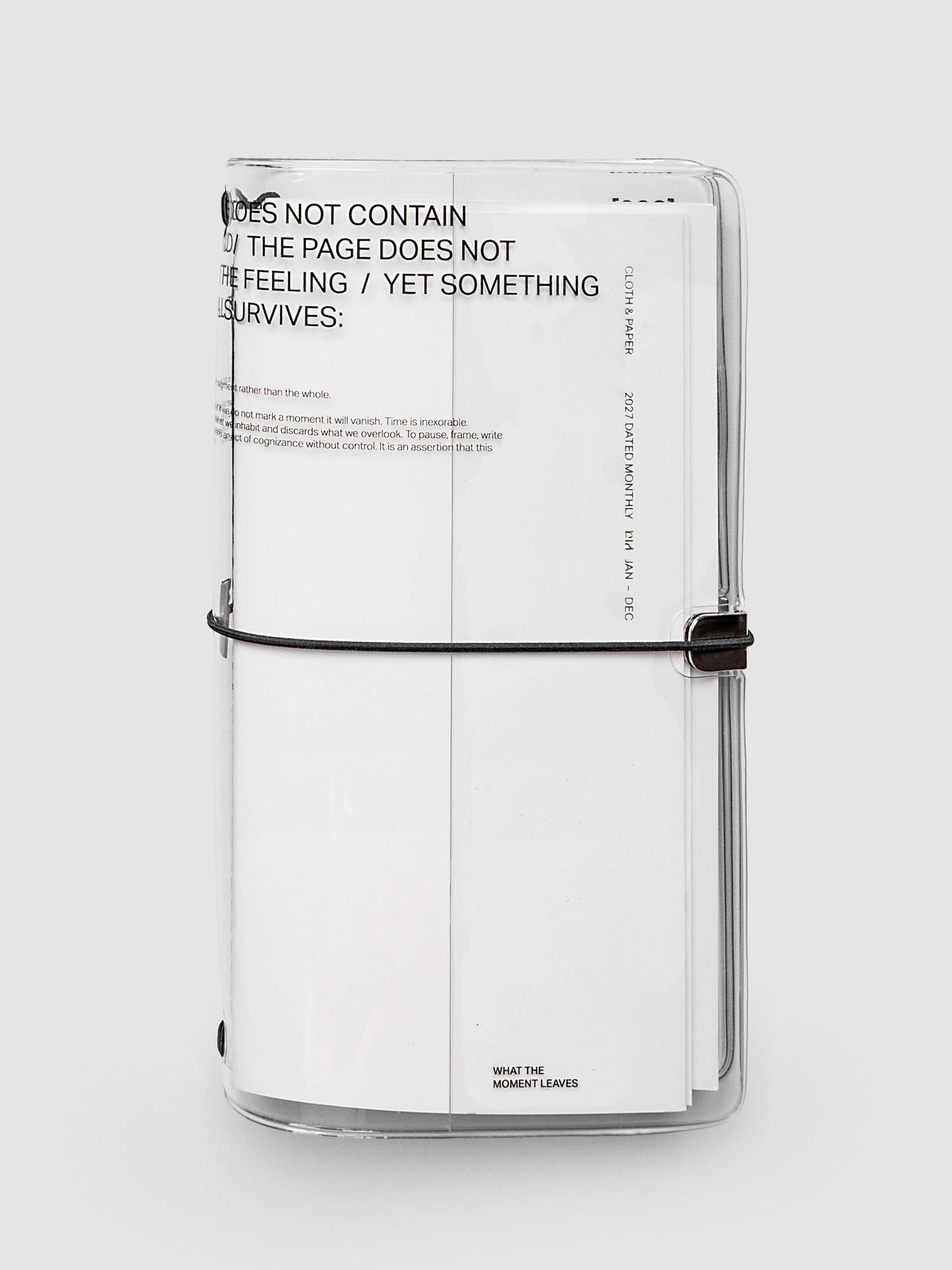 What Lasts Travel Notebook Cover in clear vinyl, A5 Slim, front exterior view, closed and upright with a white notebook loaded inside, showing typographic text on the vinyl and black bungee cord with silver metal closure
