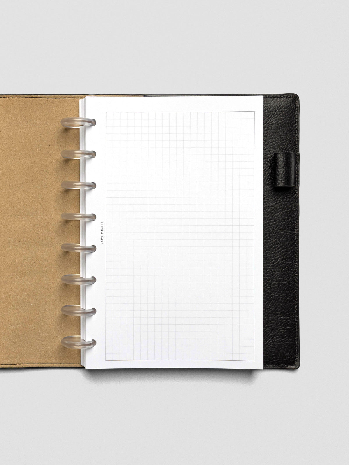A single sheet of grid paper insert with a clear border, punched with 8-disc holes, placed inside a black leather planner with tan interior lining. The layout is minimal and unmarked.