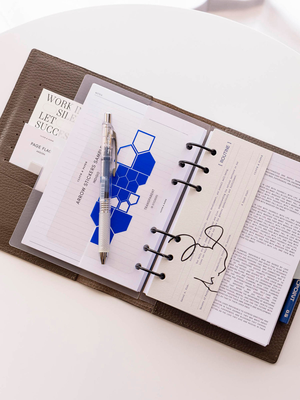 Dashboard shown in use inside a brown leather planner sitting on a white table. It is accompanied by an insert, two pens, and an indigo arrow sticker sheet.