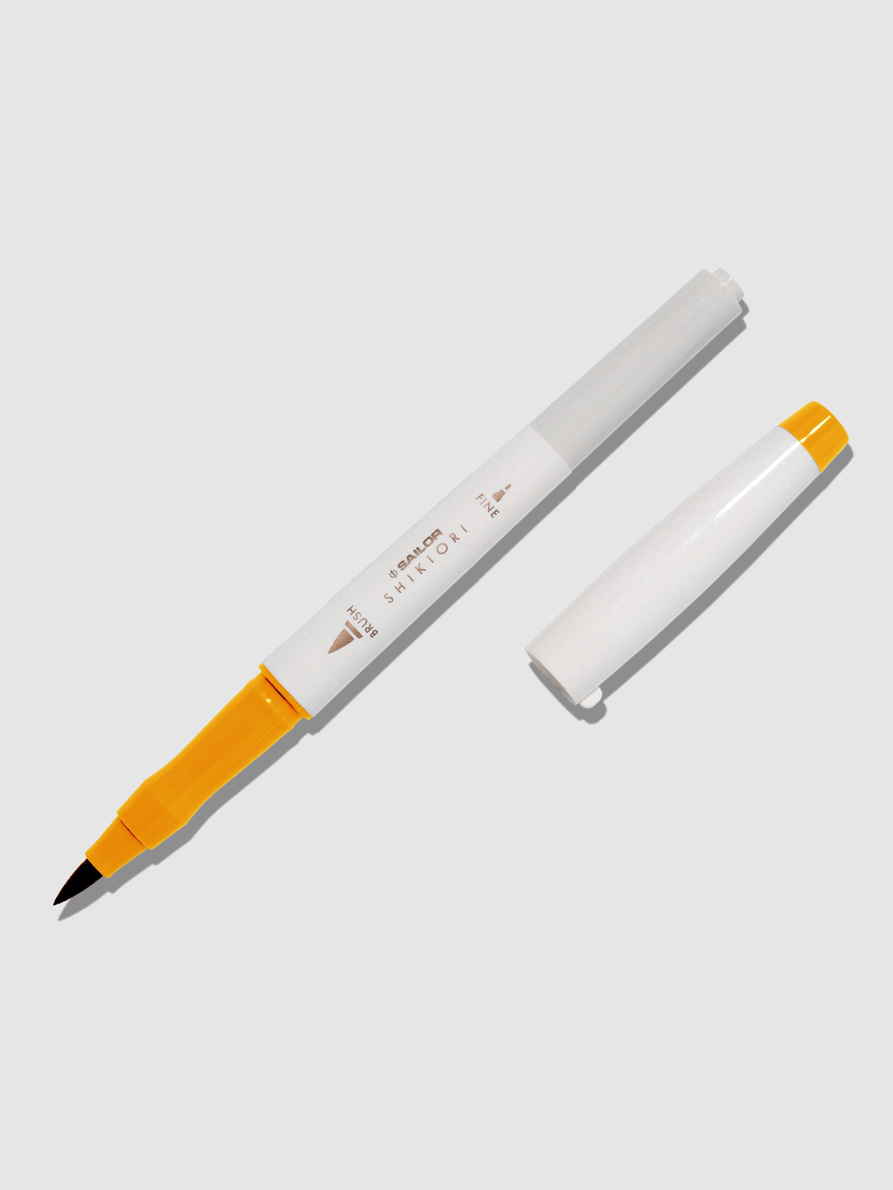 Brush pen displayed on a neutral background. It is uncapped with its brush nib exposed, and the cap lying parallel to the marker. Color pictured is Kinmokusei.
