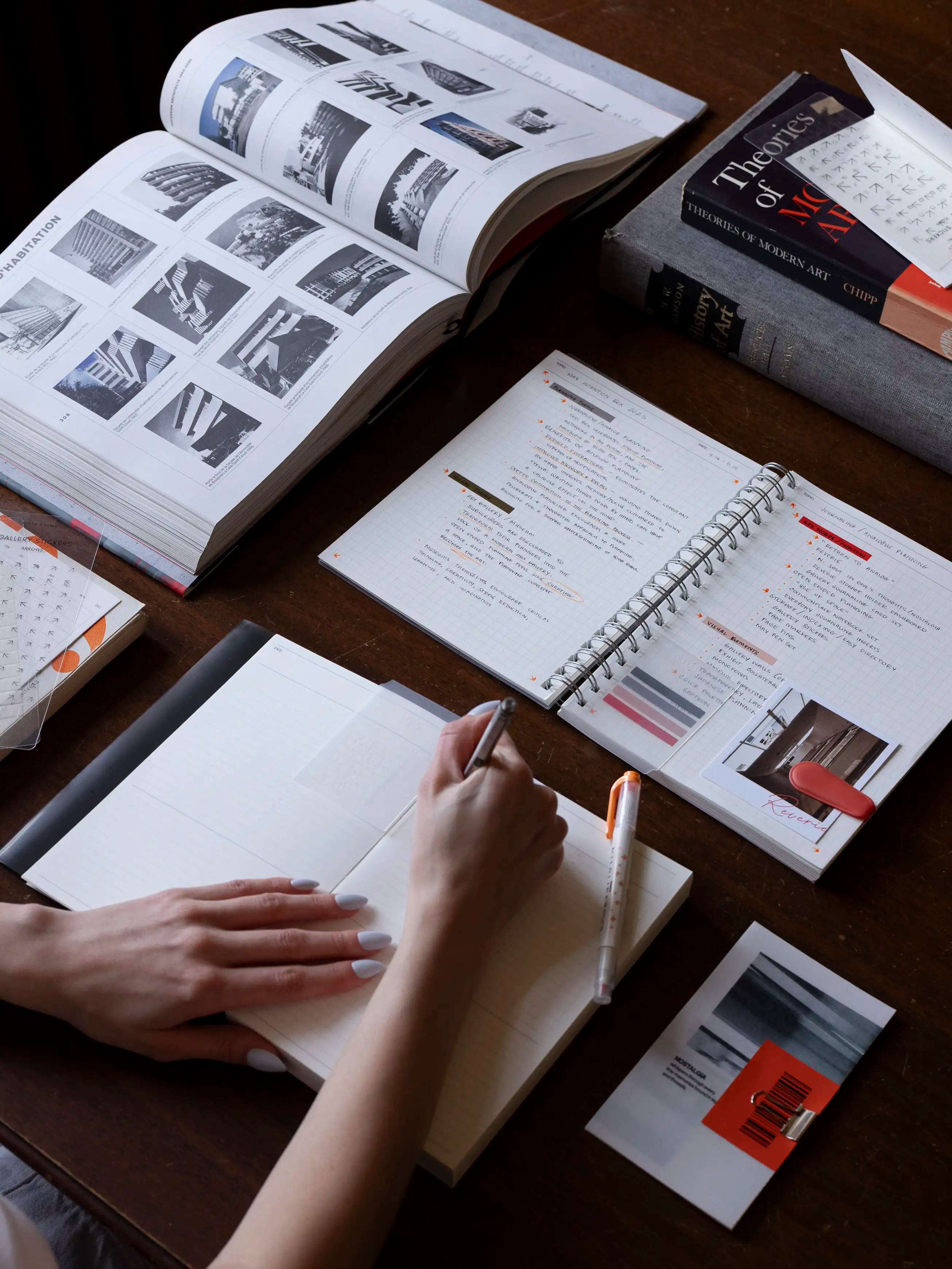 Hand writing in an open planner on a wooden desk with books, a spiral notebook of notes, and pens.