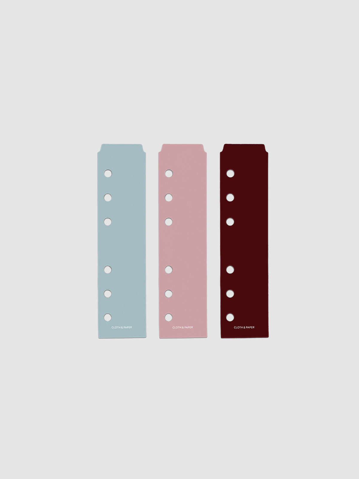 Set of three narrow page markers pre-punched for A6 planners in light blue, pink, and burgundy on a neutral background.