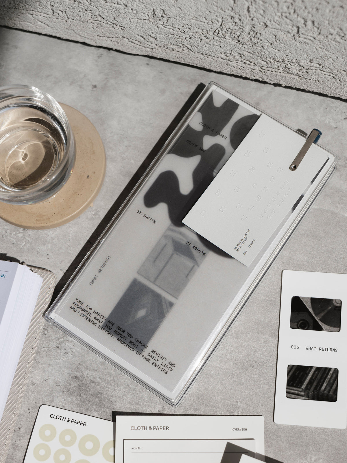 An overhead shot of a Personal Records Notebook on a grey concrete surface next to a glass of water on a wooden coaster and various stationery items, including a white bookmark and spotlight sticker sheets.