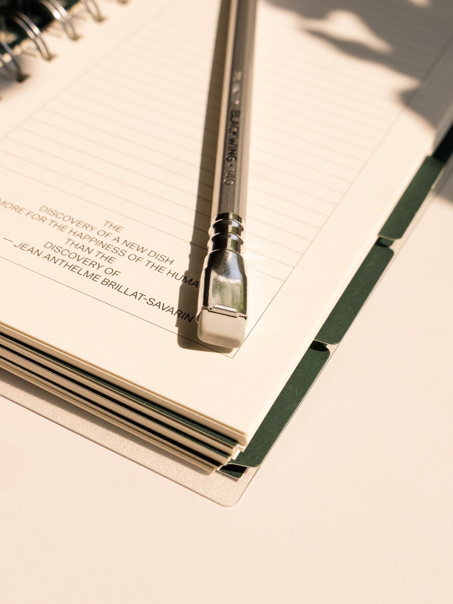 A close-up of a Silver Culinary Arts Blackwing pencil resting on the lined page of an open spiral notebook. Sunlight casts a soft shadow across the creamy paper, highlighting the pencil’s unique rectangular eraser and the printed text on the page.
