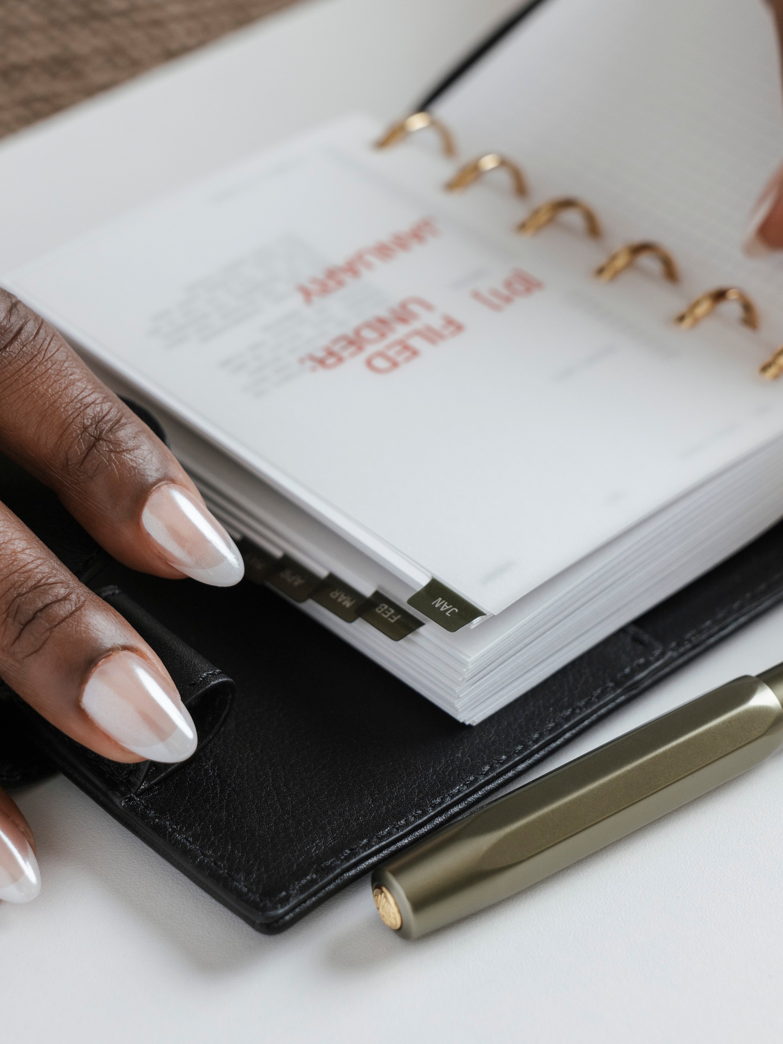 A close-up of a hand with long, white almond-shaped nails holding open a black leather ring bound planner.