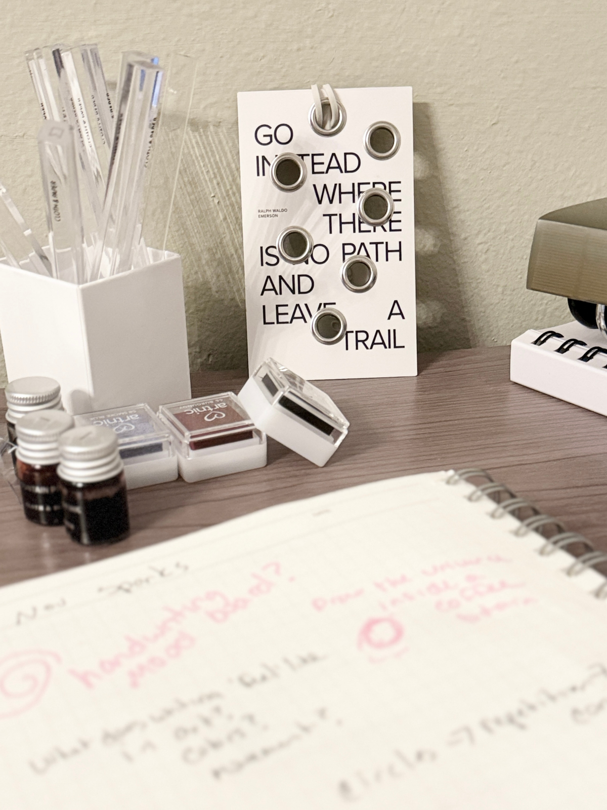 A neatly arranged desk setup featuring a motivational card that reads “Go instead where there is no path and leave a trail,” alongside a spiral notebook, ink vials, and stamp ink pads.