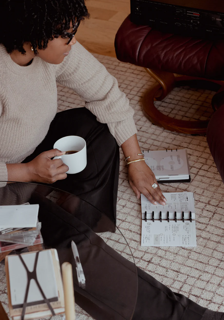 A close-up lifestyle shot of a person sitting on a rug, holding a cup of coffee while planning their day using their April 2026 Intention Box items.