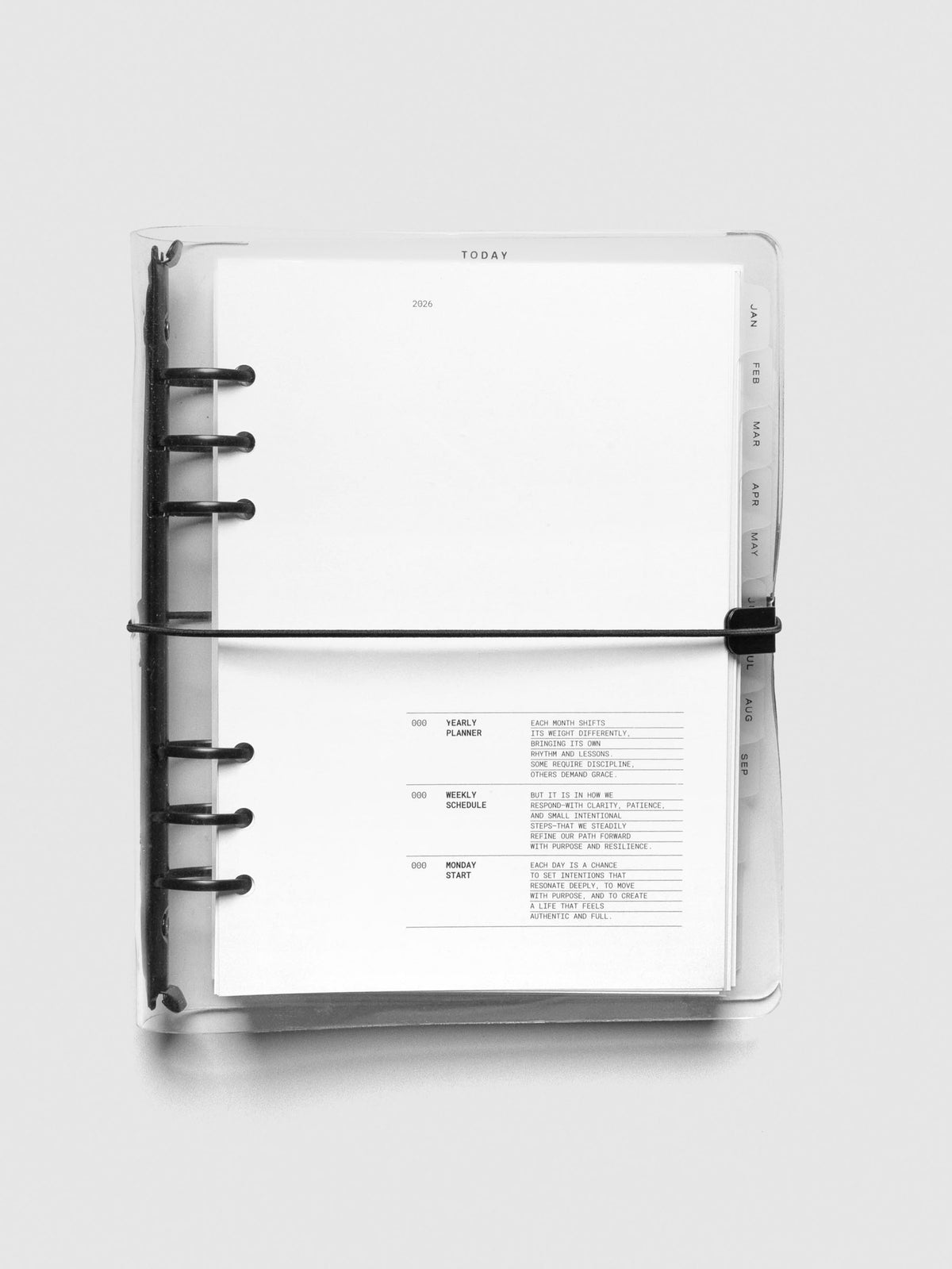 Top-down view of an A5 6-ring planner with a clear cover and black discs, showing the 2026 title page and the first planning page with intro text. Monthly tab dividers are visible along the right edge.