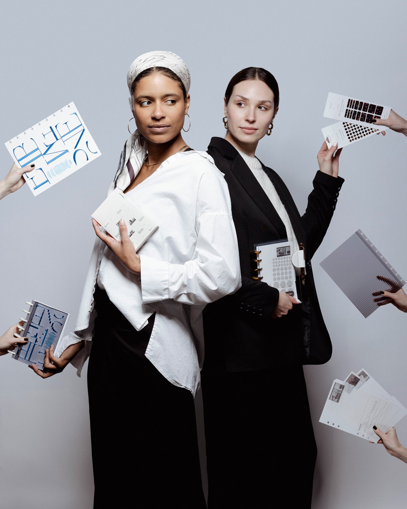 Two models are standing against a light gray background holding planner items from the Front Row Collection as hands coming in from the left and right are attempting to hand them more of these planner essentials.