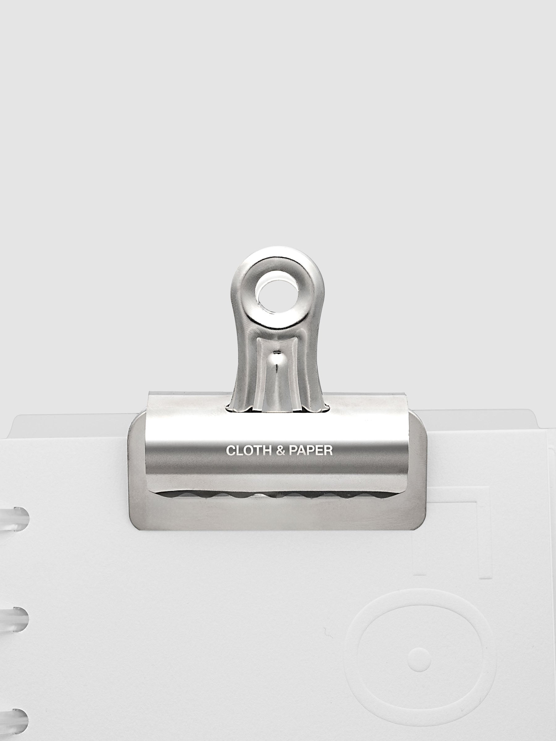 Silver metal notebook clip with Cloth & Paper logo etched onto the surface shown clipped onto a notebook.