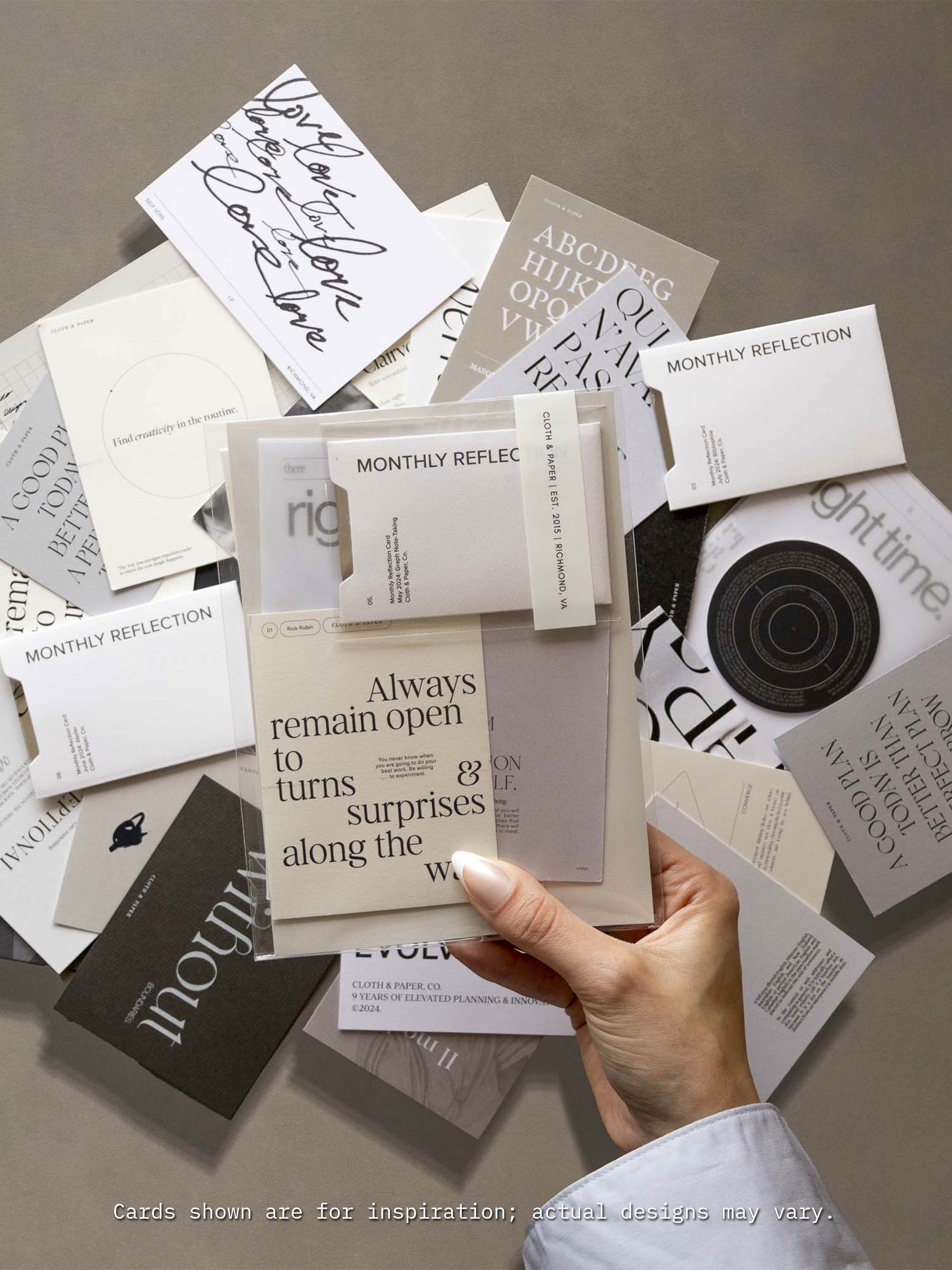 A hand holds a neutral-toned journaling card that reads ‘Always remain open to turns & surprises along the way’ above a flat lay of assorted Cloth & Paper journaling cards in minimalist fonts and designs, showcasing the variety included in the Assorted Journaling Card Set.