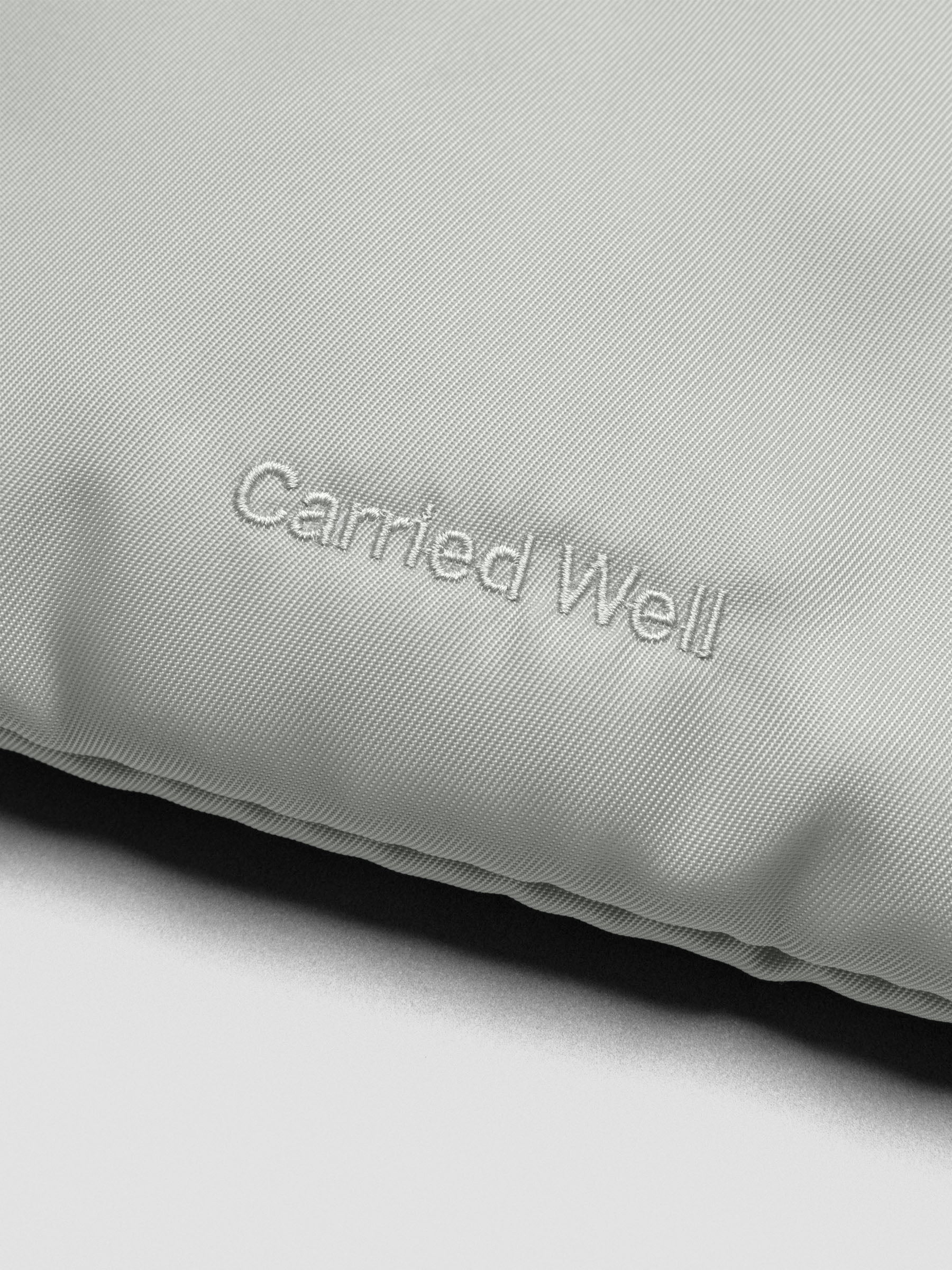 Close up of embroidery on a light green-grey nylon, padded bag reading "Carried Well".