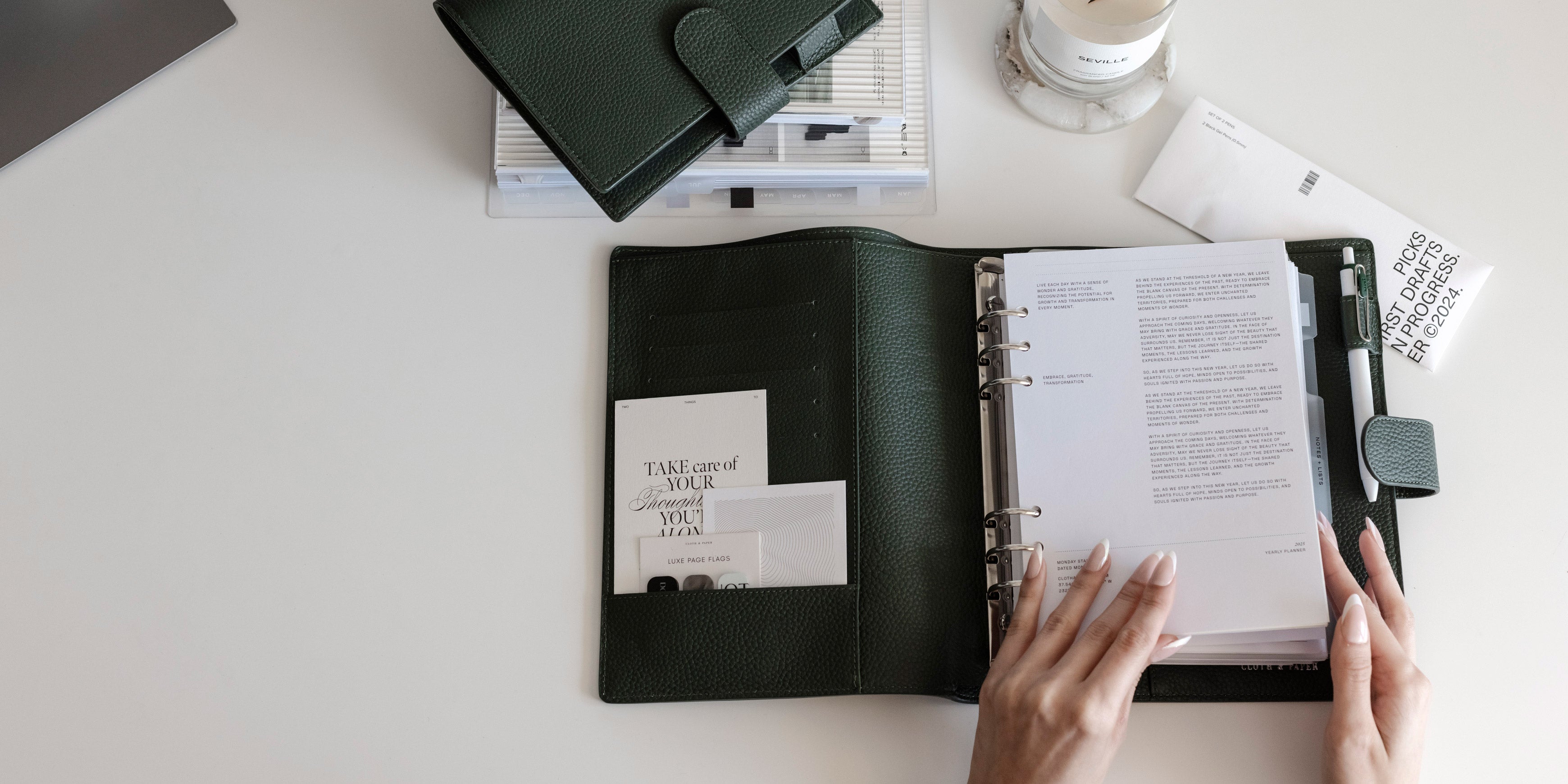 A Foundations Leather 6-Ring Agenda in A5 is opened on a white desk.  Two hands are beginning to flip through the pages inside.  Above is a Foundations Leather 6-Ring Agenda in Personal with other surrounding planner accessories.
