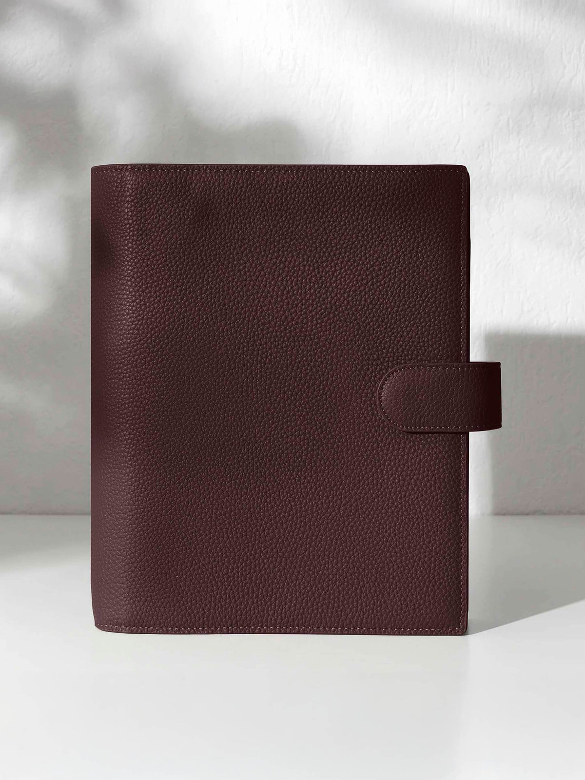 A closed dark burgundy leather planner with a closure displayed against a shadowed background.
