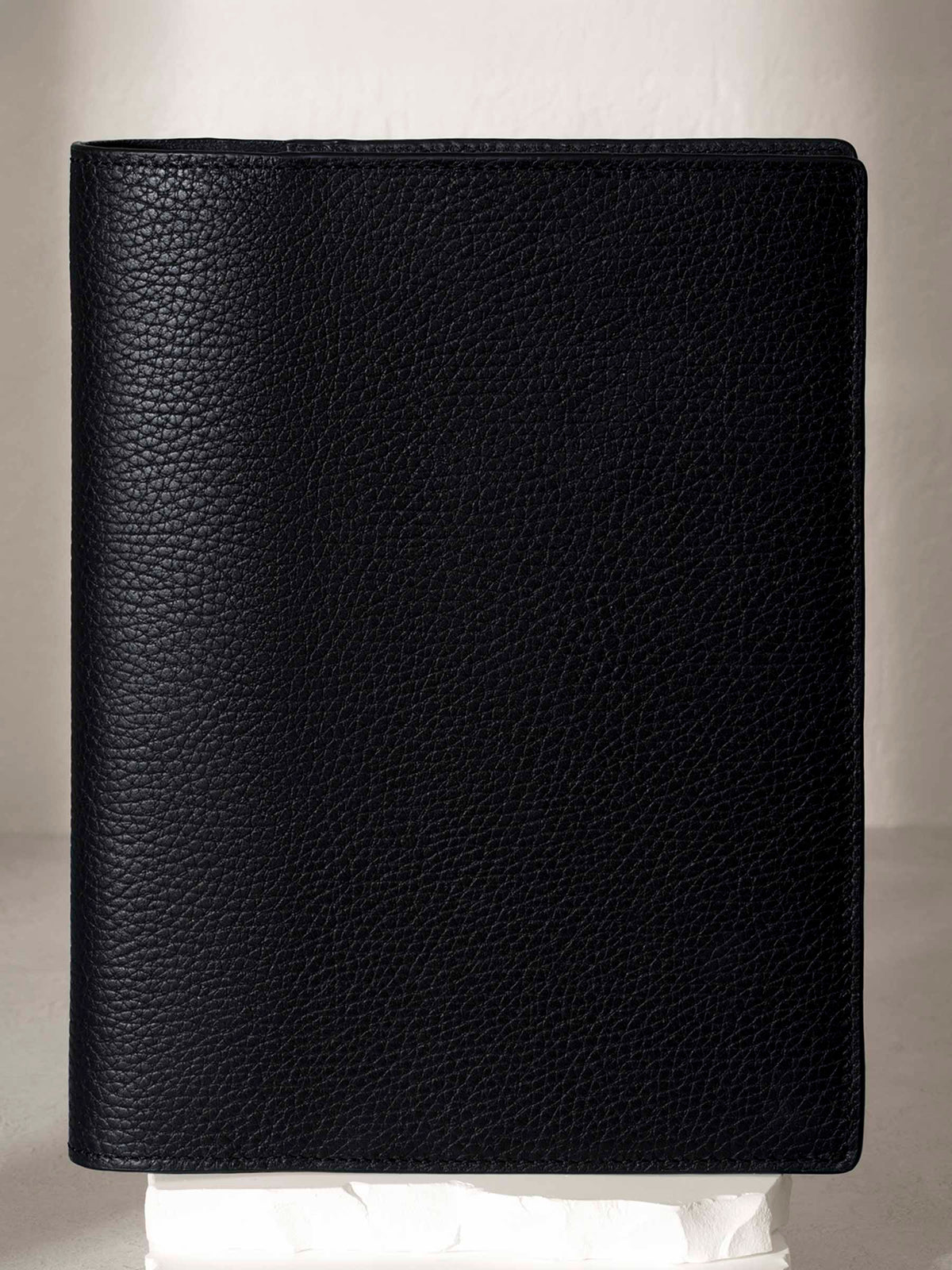 Heirloom Leather Folio, Large, Cloth and Paper. Padrillo folio displayed on a white stone pedestal. The background is a natural textured off-white material, and a spotlight behind the folio highlights its placement.