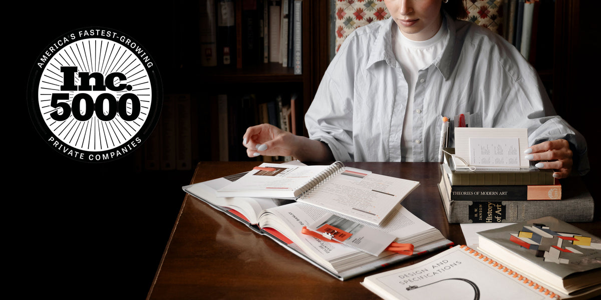 A woman is shown browsing through a pile of several books and notebooks displayed on a desk.  In the upper left corner is a logo for Inc. 5000 as Cloth & Paper was recognized for being one of America's Fastest-Growing Private Companies.