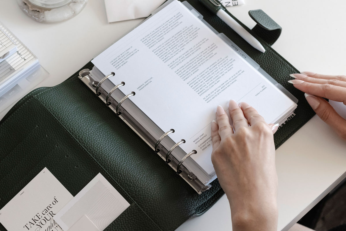 A Foundations Leather 6-Ring Agenda in A5 is opened on a white desk.  Two hands are beginning to flip through the pages inside.
