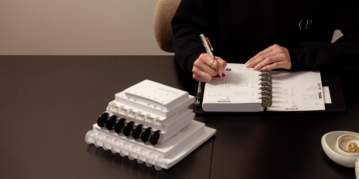 A stack of Discbound Planners and Planner Inserts.  To the right are hands of a woman who is working in an open and assembled planner featuring Speckled Planner Discs.