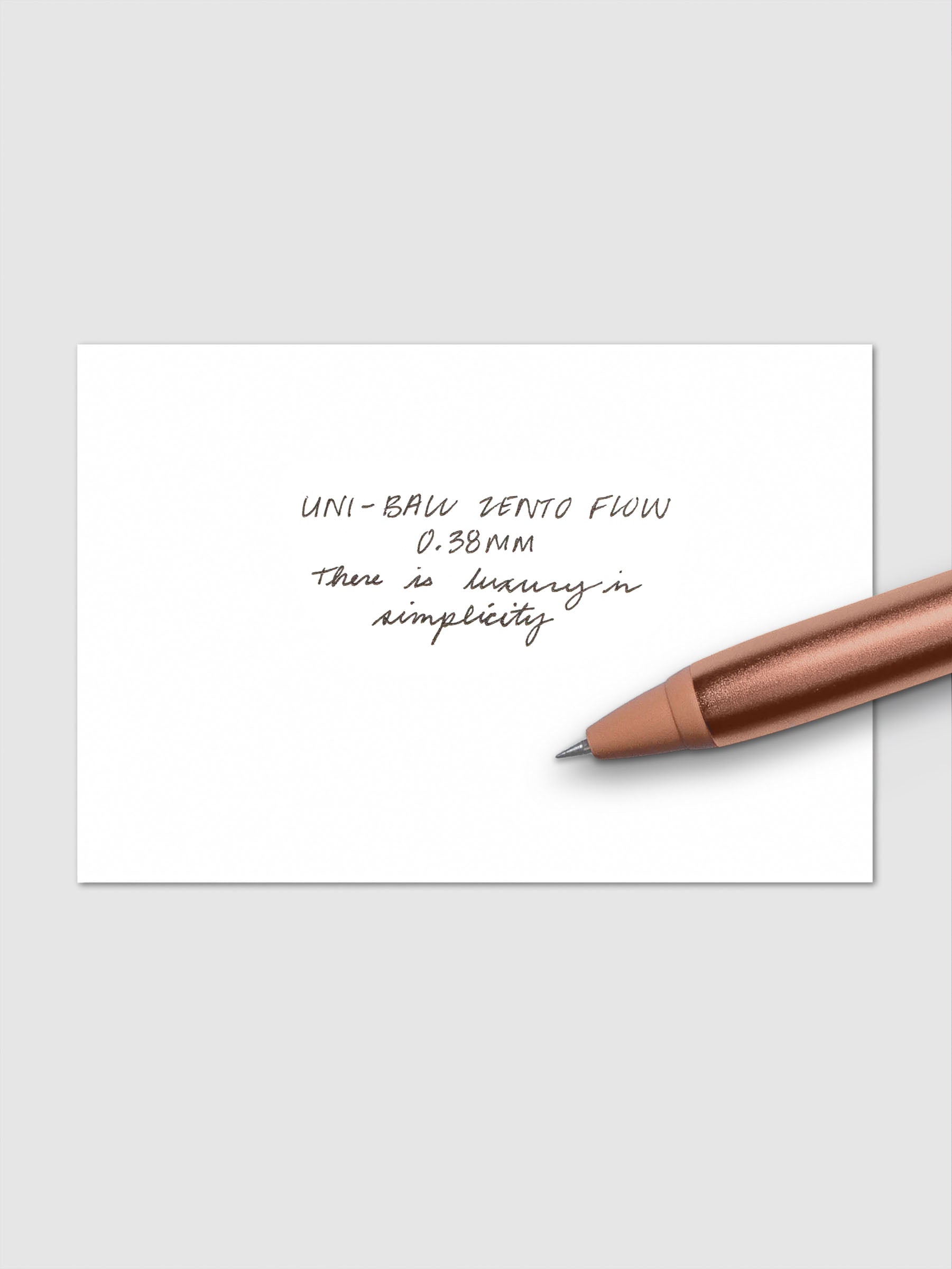 Handwritten writing sample using the Uni-ball Zento Flow Pen in a 0.38 mm nib. Shown with copper colored pen resting across paper.
